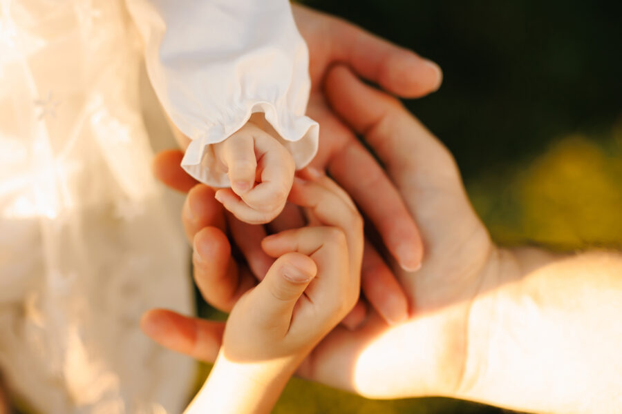 Close-up of an adult’s hands gently holding a baby’s and a child’s hands outdoors, showing a tender moment of care, connection, and support between generations in warm, natural light.
