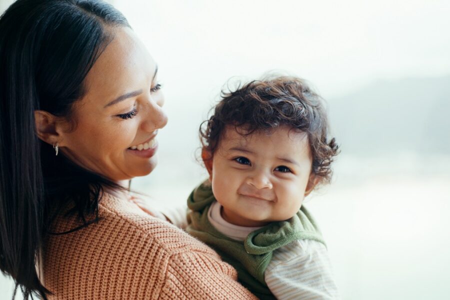 A woman smiles while holding a curly-haired baby who is looking at the camera. The background is softly lit, suggesting a bright room with windows. Both appear happy and content.
