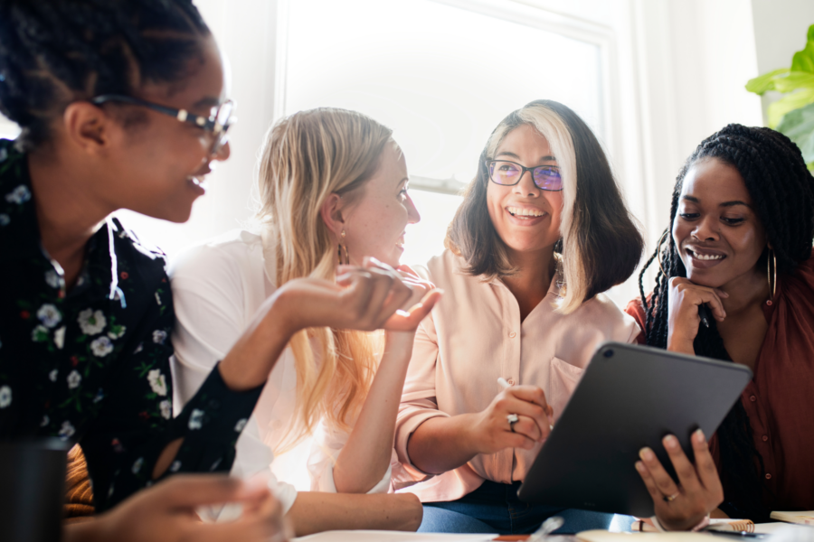 Four women sit together, smiling and chatting. One woman holds a tablet, and others listen and engage. Sunlight comes through the window behind them, creating a warm, collaborative atmosphere.