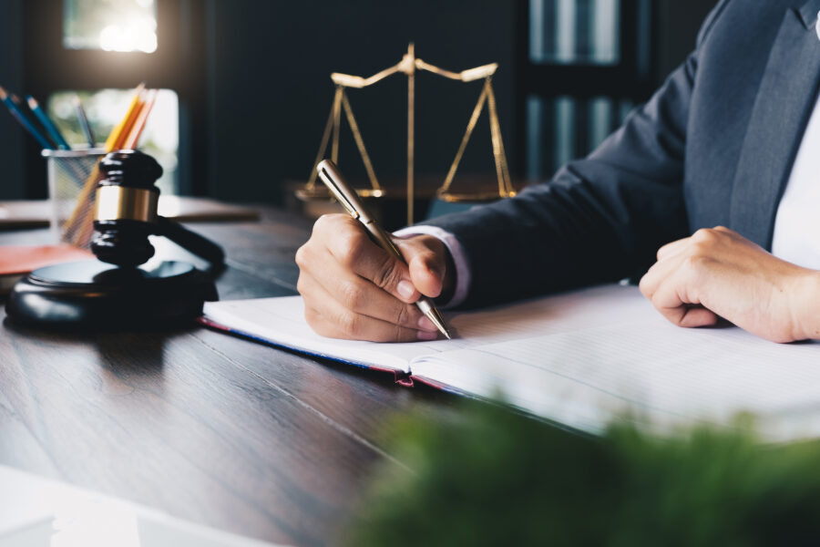 A person in a suit writes in a notebook at a desk with a gavel and scales of justice, suggesting a legal or professional setting.