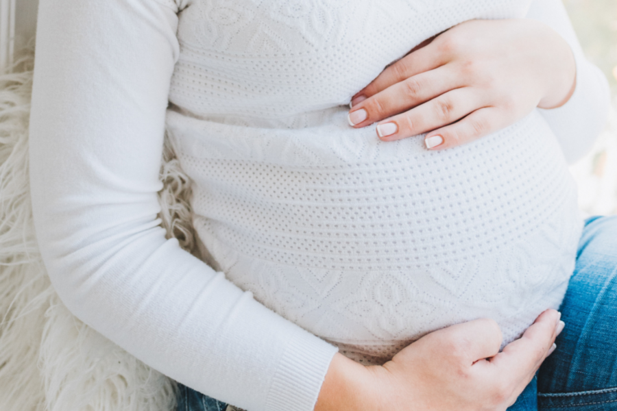 A pregnant person wearing a white textured top and blue jeans sits while gently cradling their belly with both hands.