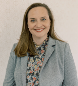 A woman with long light brown hair wearing a gray blazer and a floral blouse smiles at the camera against a light patterned background.