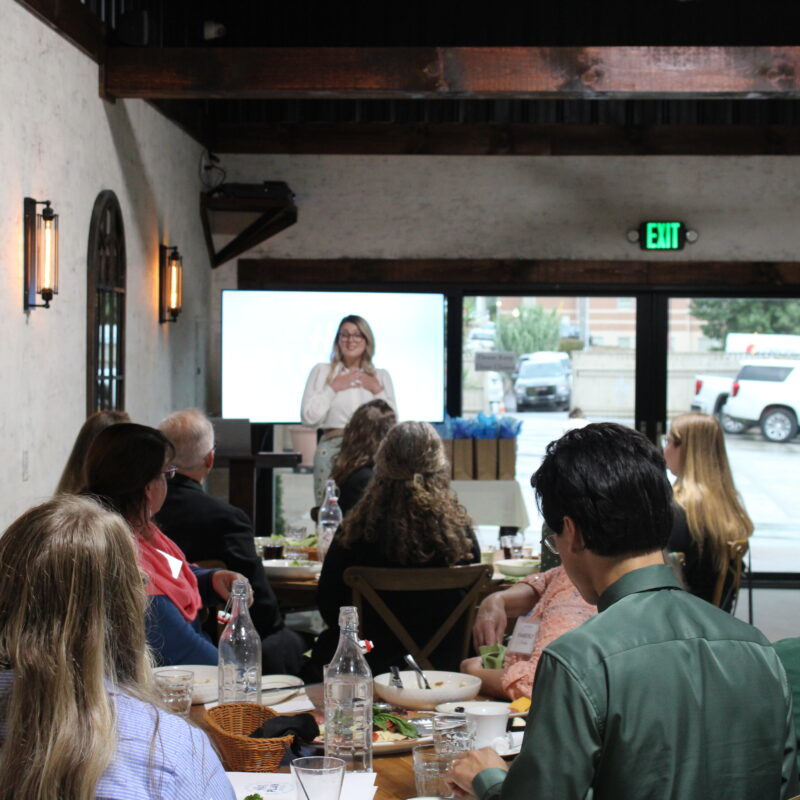 A woman stands and speaks at the front of a rustic indoor event space, addressing a seated audience. Attendees are dining at tables while listening, and large windows show parked cars outside.