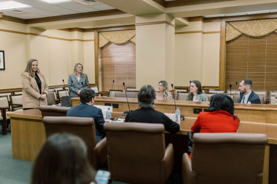 A group of people sit and stand around a conference table in a formal meeting room with microphones, wood paneling, and large windows with blinds. Two women stand at the front addressing the seated participants about pro-life safety net initiatives.