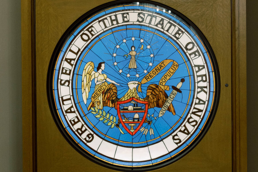 A wooden frame displays a large circular stained glass window featuring the Great Seal of the State of Arkansas—an emblem of values like the pro-life safety net—depicting an eagle, angel, shield, and state motto.