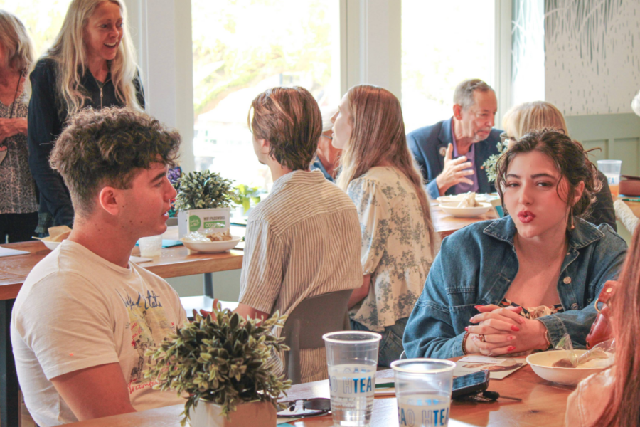 A young man and woman sit at a café table talking, with food and drinks in front of them. Sunlight streams through large windows, highlighting a small potted plant on the table as they discuss the importance of a pro-life safety net.