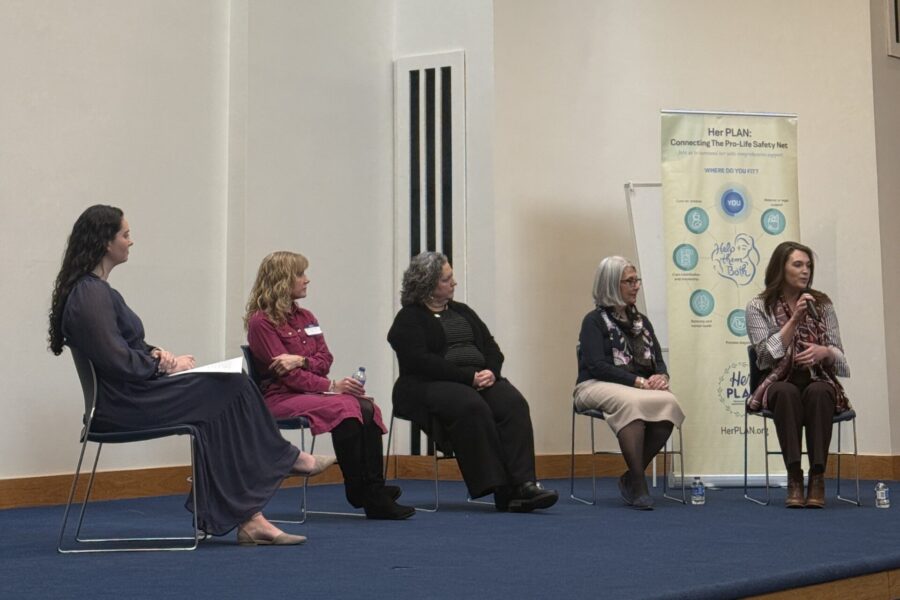 Five women sit on a stage panel, engaged in discussion about the pro-life safety net. Four are seated, one gestures while speaking. A banner with the Her PLAN logo and icons is displayed in the background, with water bottles on the floor.