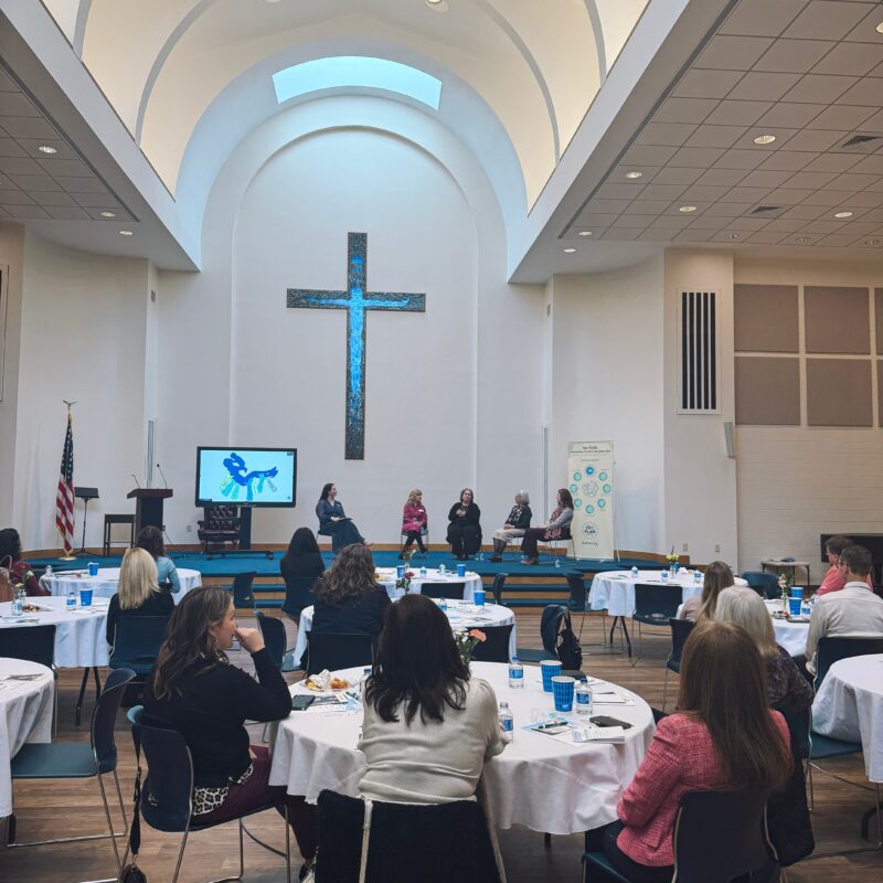People sit at round tables in a bright room with high ceilings and a prominent cross on the wall. Four panelists discuss pro-life safety net initiatives near a screen with a blue graphic, suggesting a community event or seminar.