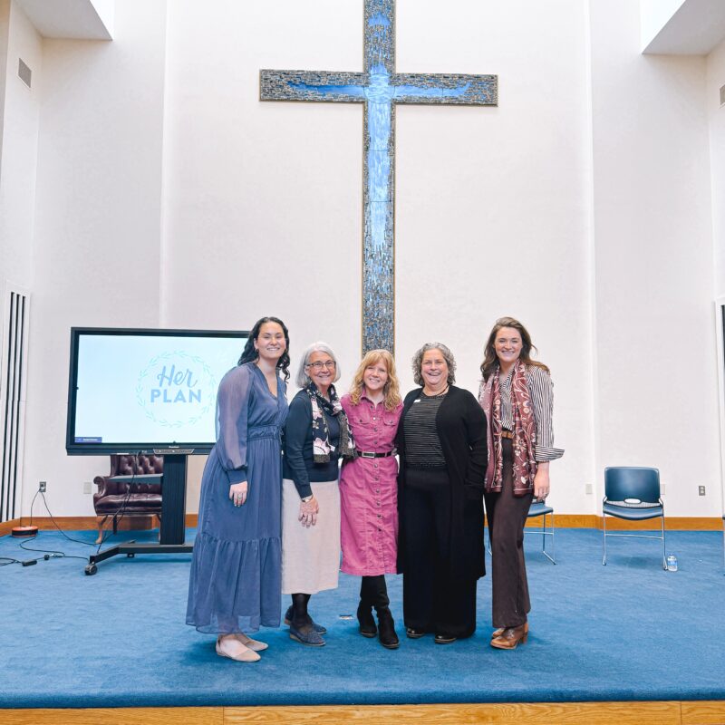 Five women stand smiling on a blue-carpeted stage in front of a large cross and an arched wall. A TV screen displaying Her Plan—a pro-life safety net initiative—is to the left, with two chairs visible in the background.