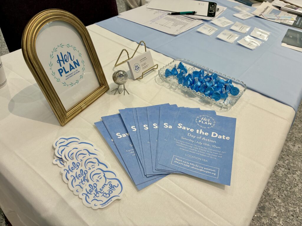 A tabletop display with a “Her Plan” framed sign, blue Save the Date flyers, stickers, business cards, and blue-wrapped chocolates highlights resources from a pro-life safety net—all neatly arranged on a white tablecloth.