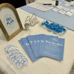 A tabletop display with a “Her Plan” framed sign, blue Save the Date flyers, stickers, business cards, and blue-wrapped chocolates highlights resources from a pro-life safety net—all neatly arranged on a white tablecloth.