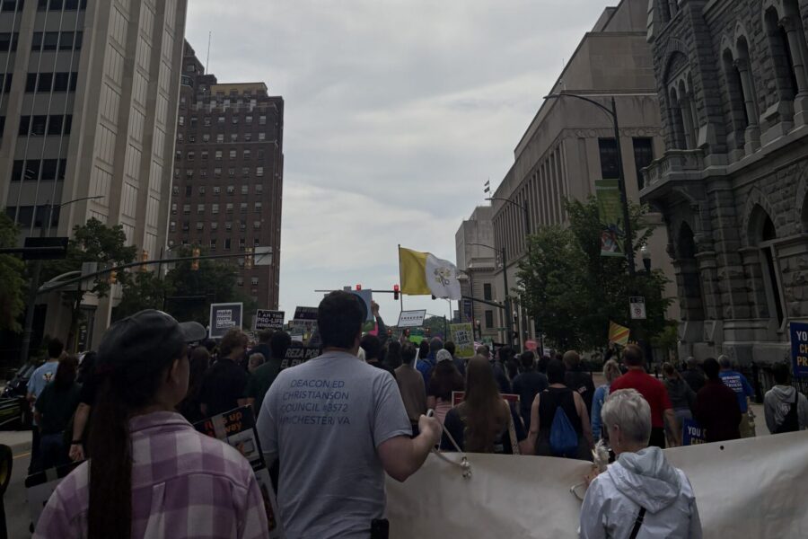 A crowd of people marches down a city street between tall buildings under a cloudy sky, holding signs and banners calling for a stronger pro-life safety net during a protest.