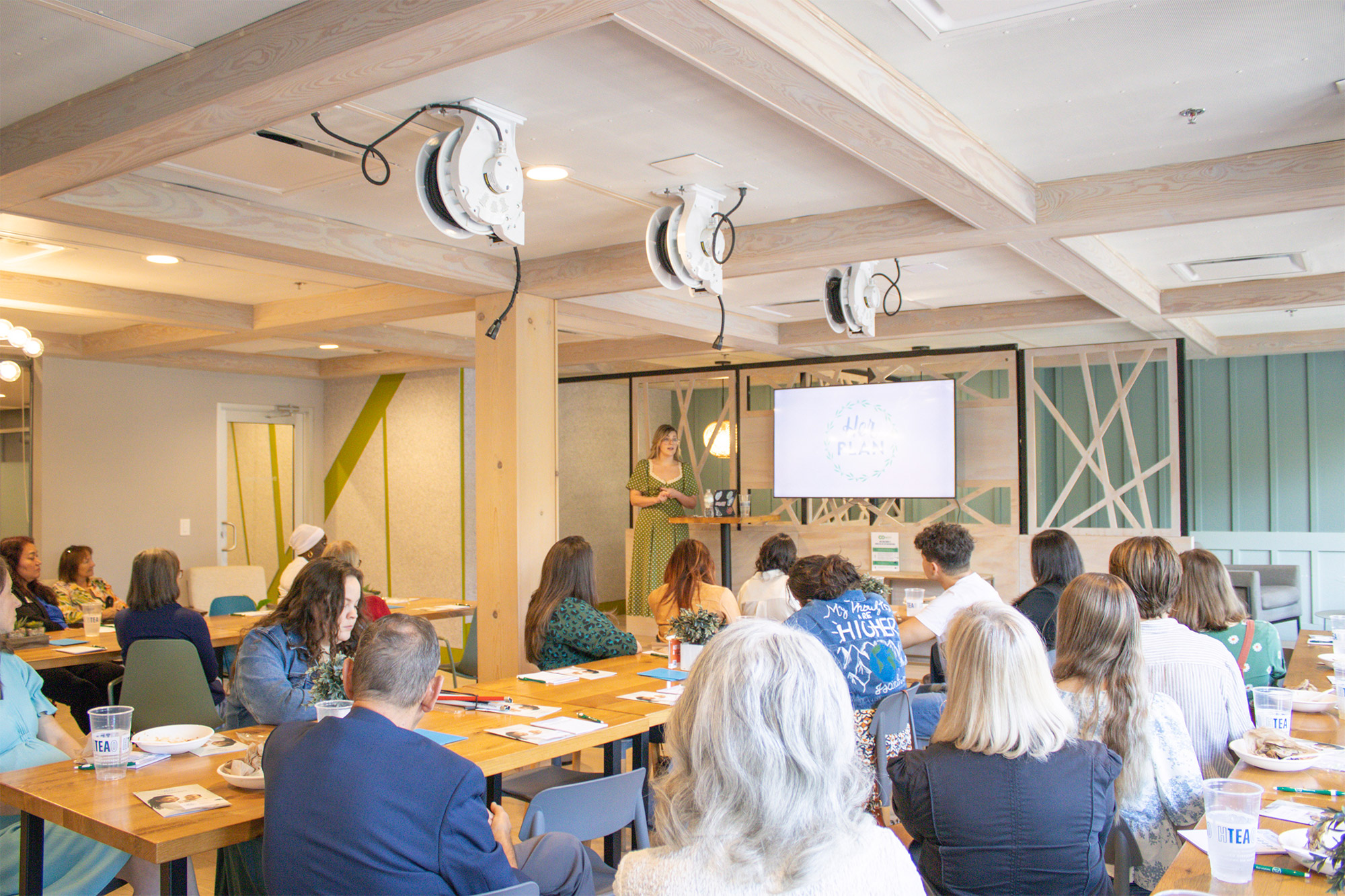 A group of people sit at tables in a bright conference room, facing a woman giving a presentation in front of a screen displaying a circular logo. Papers, drinks, and food are on the tables.