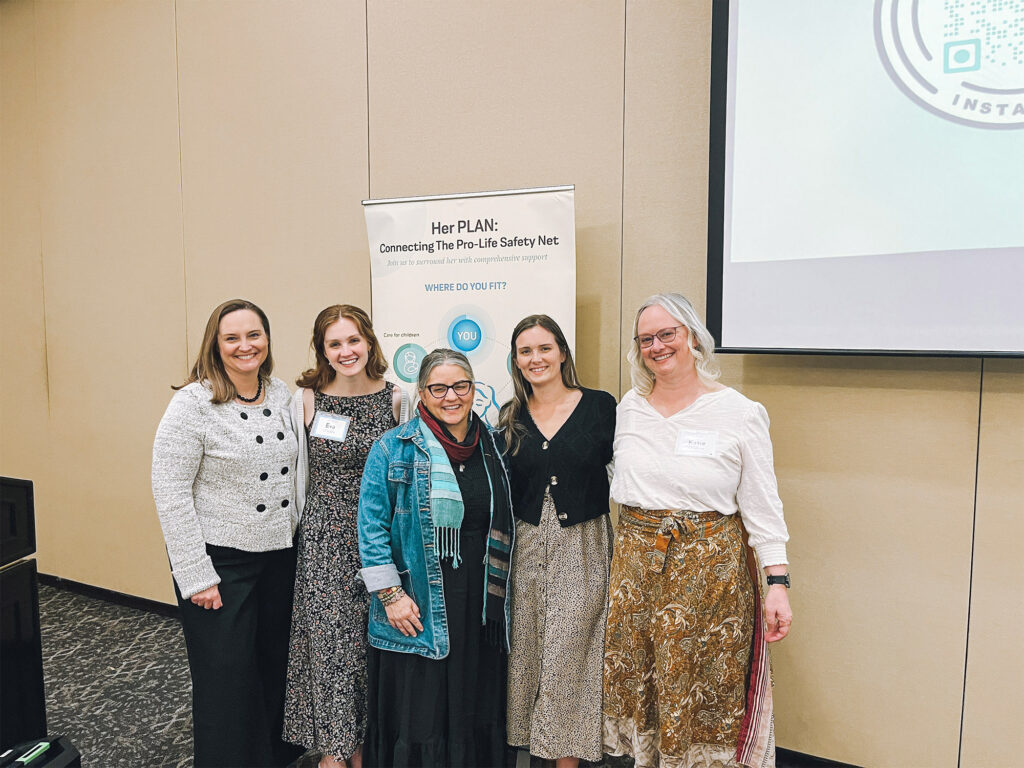 Five women smiling and standing together indoors in front of a beige wall and a banner that reads Her PLAN: Connecting The Pro-Life Safety Net. A projector screen is visible on the right side.
