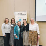 Five women smiling and standing together indoors in front of a beige wall and a banner that reads Her PLAN: Connecting The Pro-Life Safety Net. A projector screen is visible on the right side.