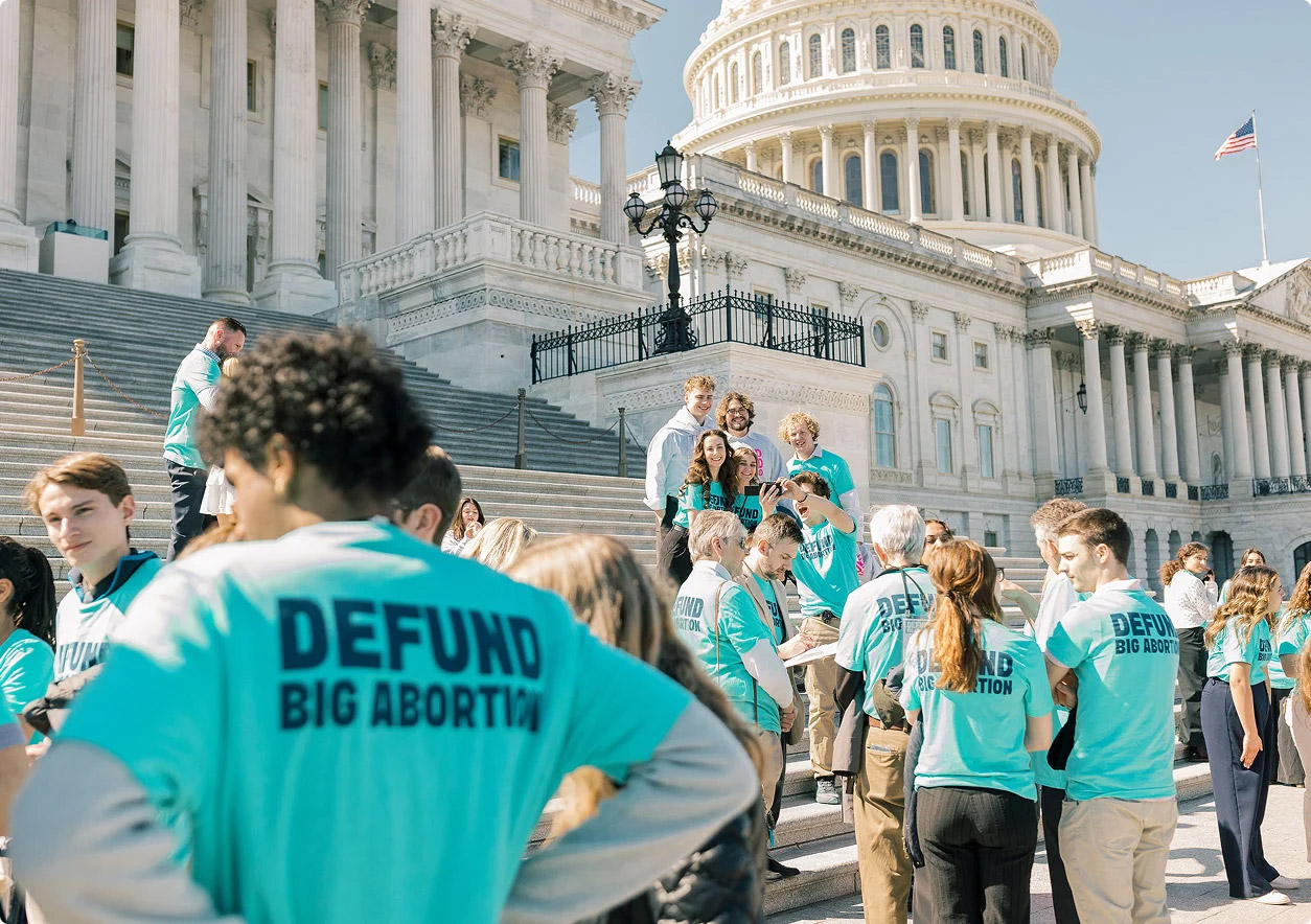A group of people wearing matching turquoise shirts that read DEFUND BIG ABORTION gather and talk on the steps outside a large government building with columns and a dome.