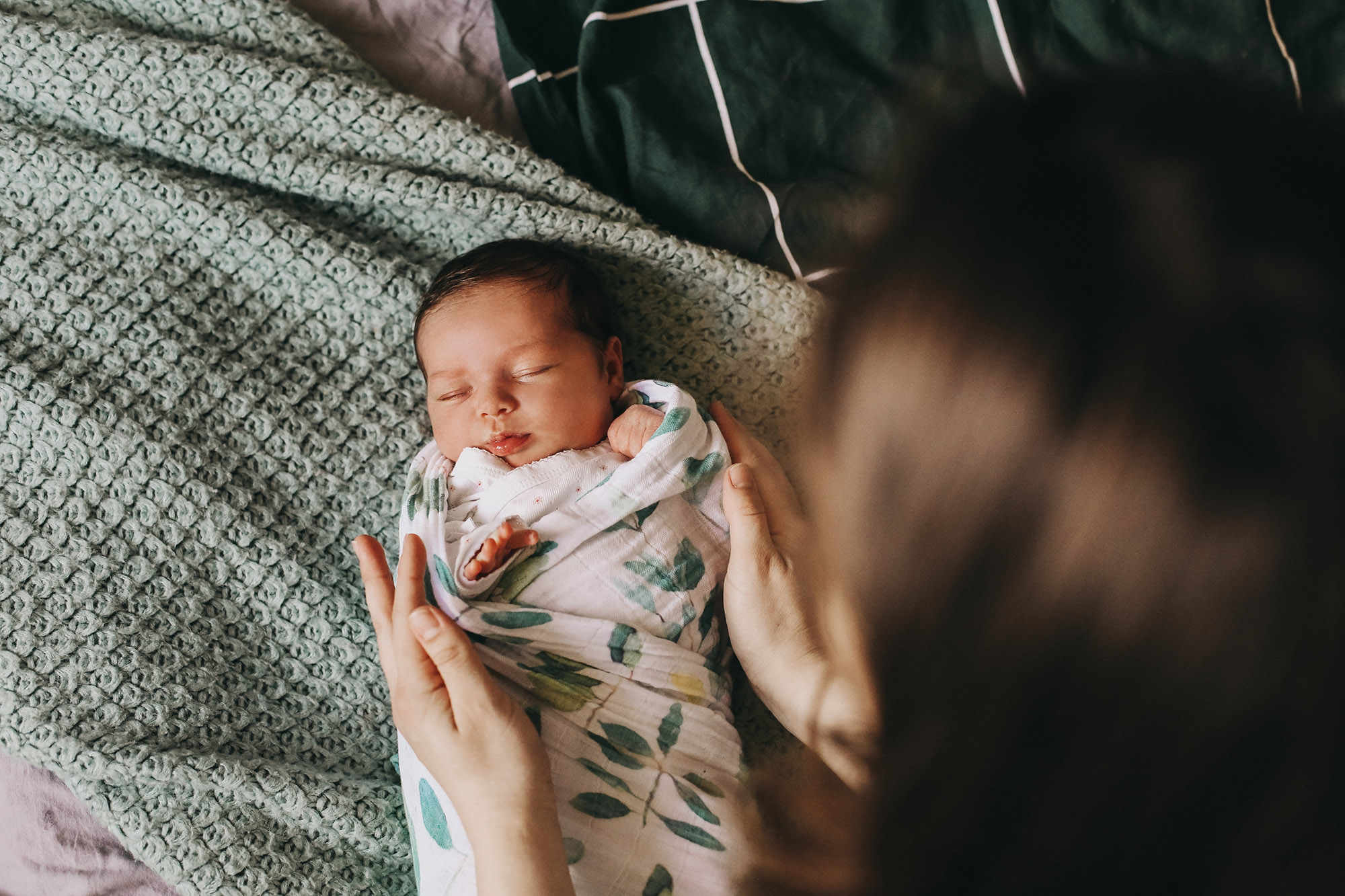 A newborn baby wrapped in a patterned blanket sleeps on a bed while an adult gently places their hands nearby, creating a caring and protective scene.