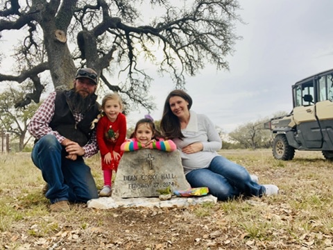 A man, two young girls, and a pregnant woman—symbolizing the importance of a pro-life safety net—sit together outdoors by a memorial stone that reads Indian Rocky Hall, February 20, 2021. Trees and an off-road vehicle are in the background.