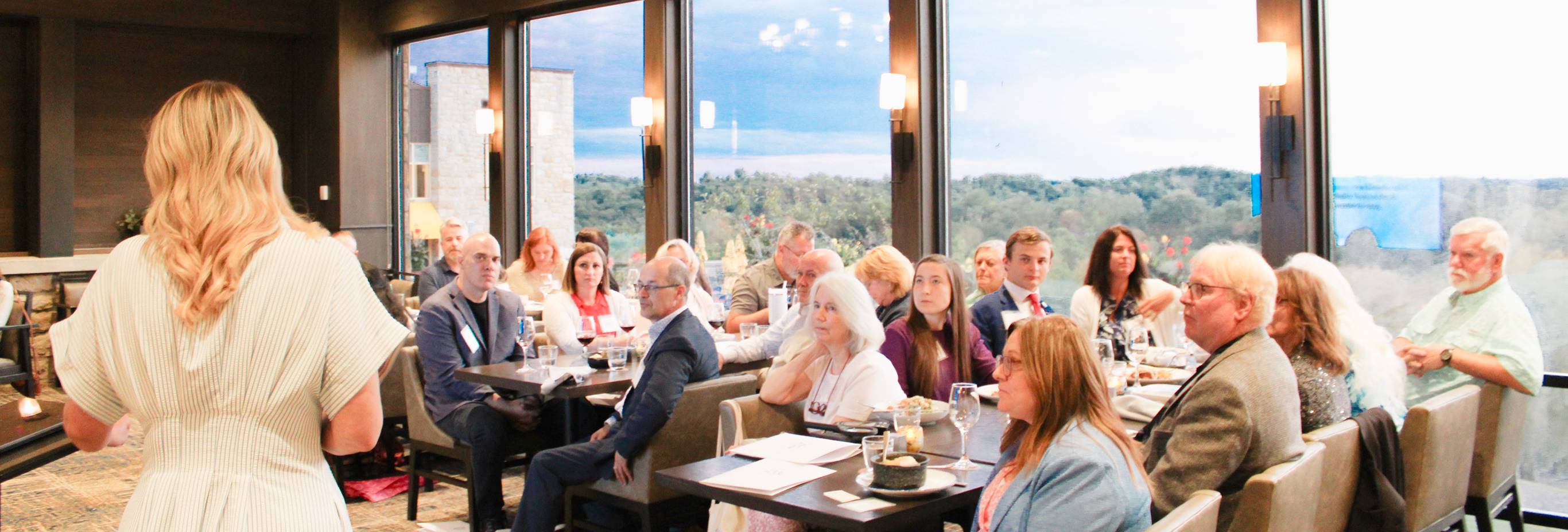 A woman stands and speaks to a group of people seated at tables in a well-lit room with large windows and a scenic outdoor view. The audience is attentively listening to her presentation.
