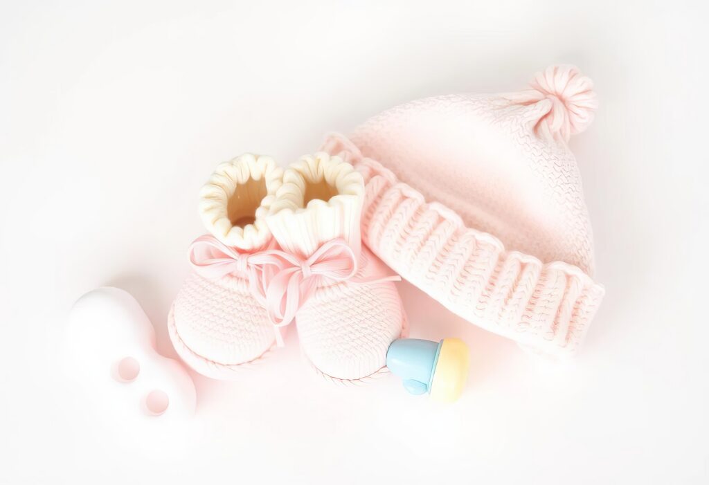 Soft pink baby hat and booties with bows, a yellow and blue pacifier, and a white plastic rattle are arranged on a white surface.