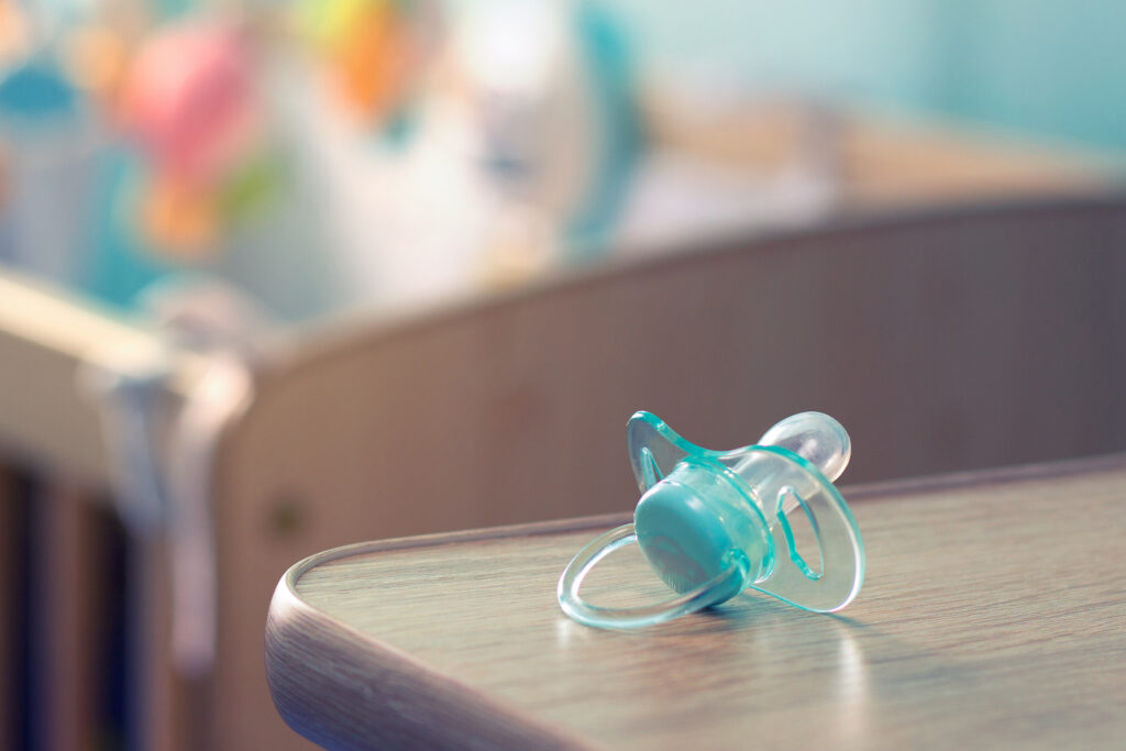 A translucent blue pacifier rests on the edge of a wooden table, with a blurred wooden crib and soft, pastel-colored background in a nursery room.