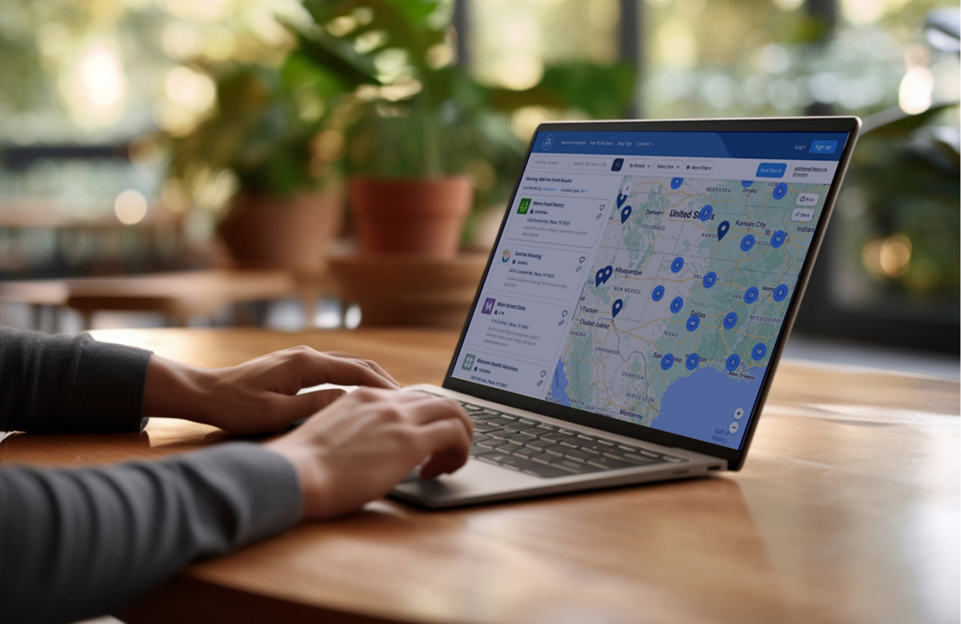 Person using a laptop at a wooden table, viewing a map with location pins and a list of places on the screen. Green plants and large windows are visible in the blurred background.