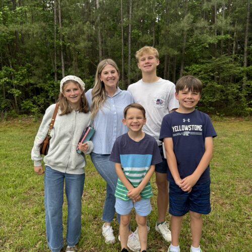 Five smiling children stand together on green grass with trees in the background. Reflecting a pro-life safety net, they wear casual clothes; one holds a book and wears a headband. The group appears happy and relaxed outdoors.