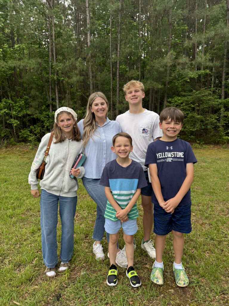 A woman and four children stand smiling on green grass with a forest background. The children wear casual clothes; one girl holds notebooks, and the boys wear shorts and T-shirts. Everyone looks relaxed and happy outdoors.
