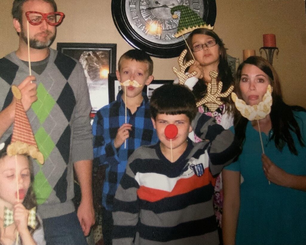 Six people, including adults and children, pose for a playful photo holding up holiday-themed props like fake beards, reindeer antlers, glasses, hats, and a red nose. A large wall clock is visible in the background.