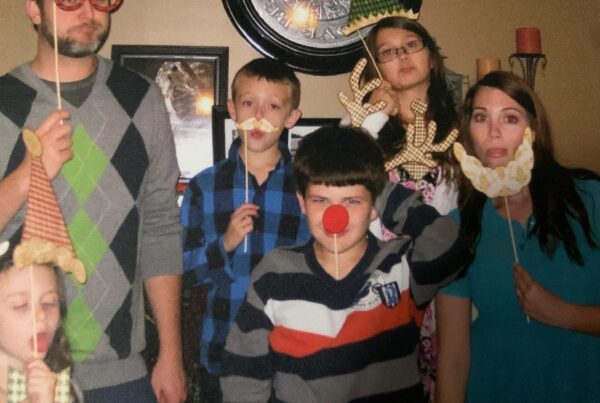 A group of five people, including children and adults, pose indoors holding playful props like fake glasses, hats, a red nose, and beards. They stand in front of a wall clock, showing silly and serious faces—highlighting the joy found within a pro-life safety net.