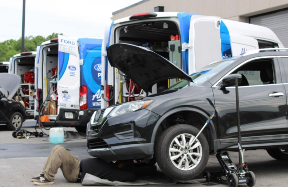 A mechanic lies under a black SUV with its hood open, performing maintenance. Two service vans with open doors and visible equipment are parked nearby in an outdoor area.