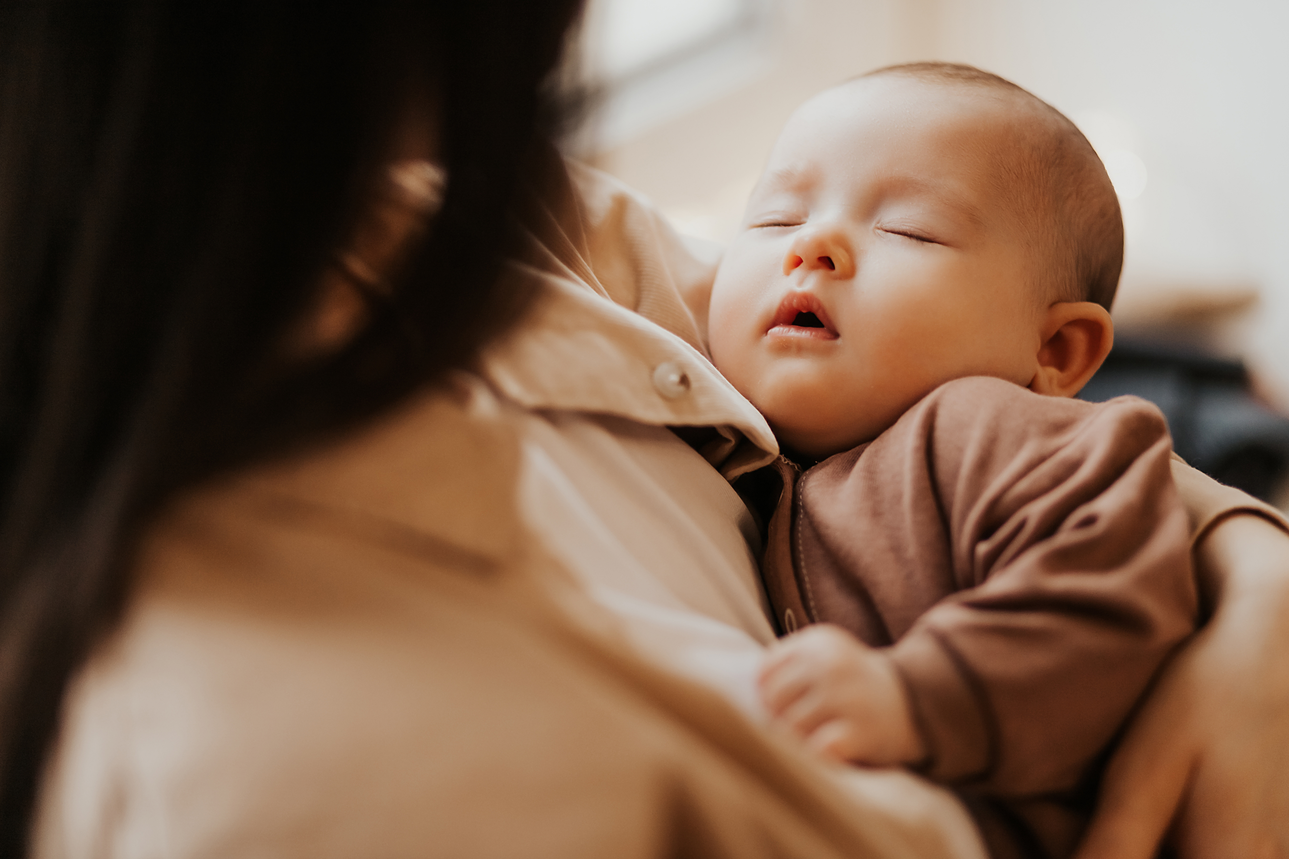 A baby sleeps peacefully in someones arms, dressed in a soft brown outfit. The person holding the baby is seen from behind, providing a sense of comfort and care. The background is softly blurred.