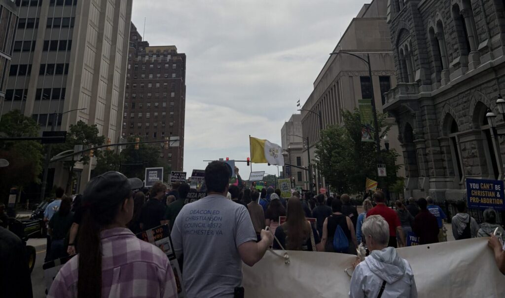 A large group of people march down a city street holding banners and signs, some with slogans about abortion rights. Tall buildings line both sides, and a yellow flag is visible in the distance.