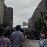A large group of people march down a city street holding banners and signs, some with slogans about abortion rights. Tall buildings line both sides, and a yellow flag is visible in the distance.
