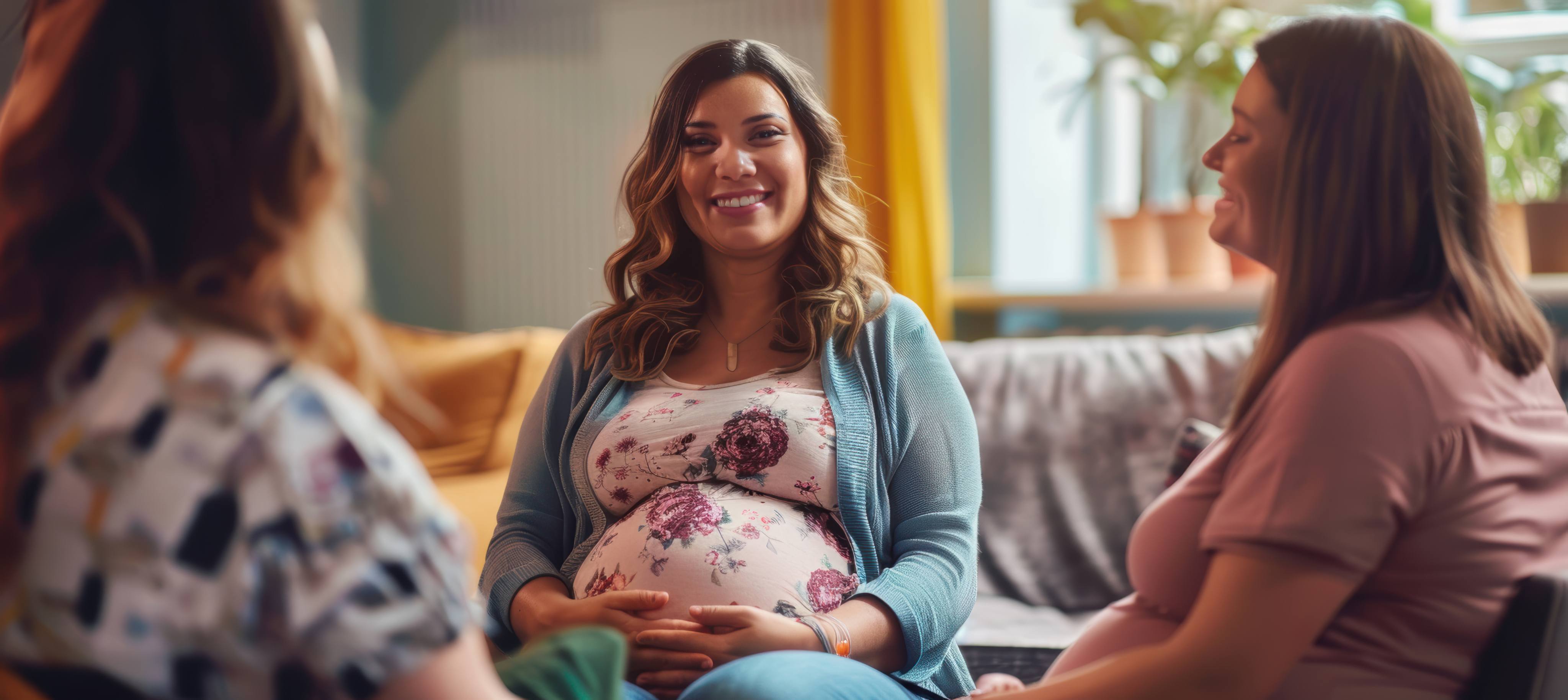 Three pregnant women sit together on a couch in a cozy living room, smiling and talking. Sunlight streams through a window with plants on the sill, creating a warm and supportive atmosphere.