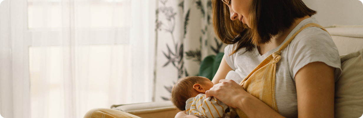 A woman sits on a couch by a window, breastfeeding her baby. The scene is softly lit by natural light, and the mother looks lovingly at her child. A patterned curtain is visible in the background.