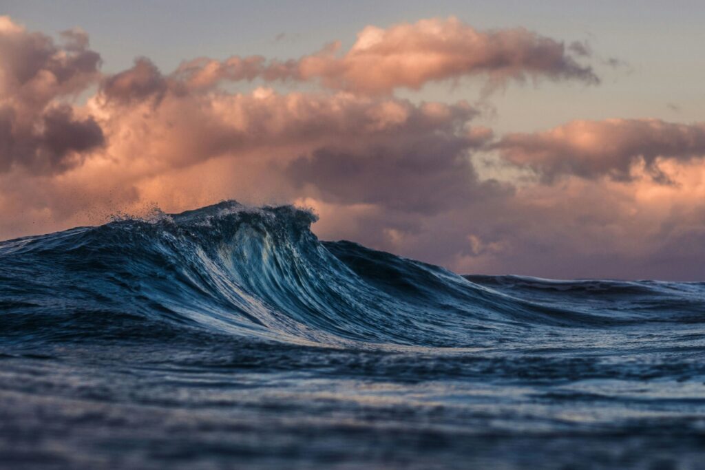 A close-up of a blue ocean wave with dramatic clouds in the sky, illuminated by the soft pink and orange hues of sunset or sunrise.