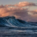 A close-up of a blue ocean wave with dramatic clouds in the sky, illuminated by the soft pink and orange hues of sunset or sunrise.