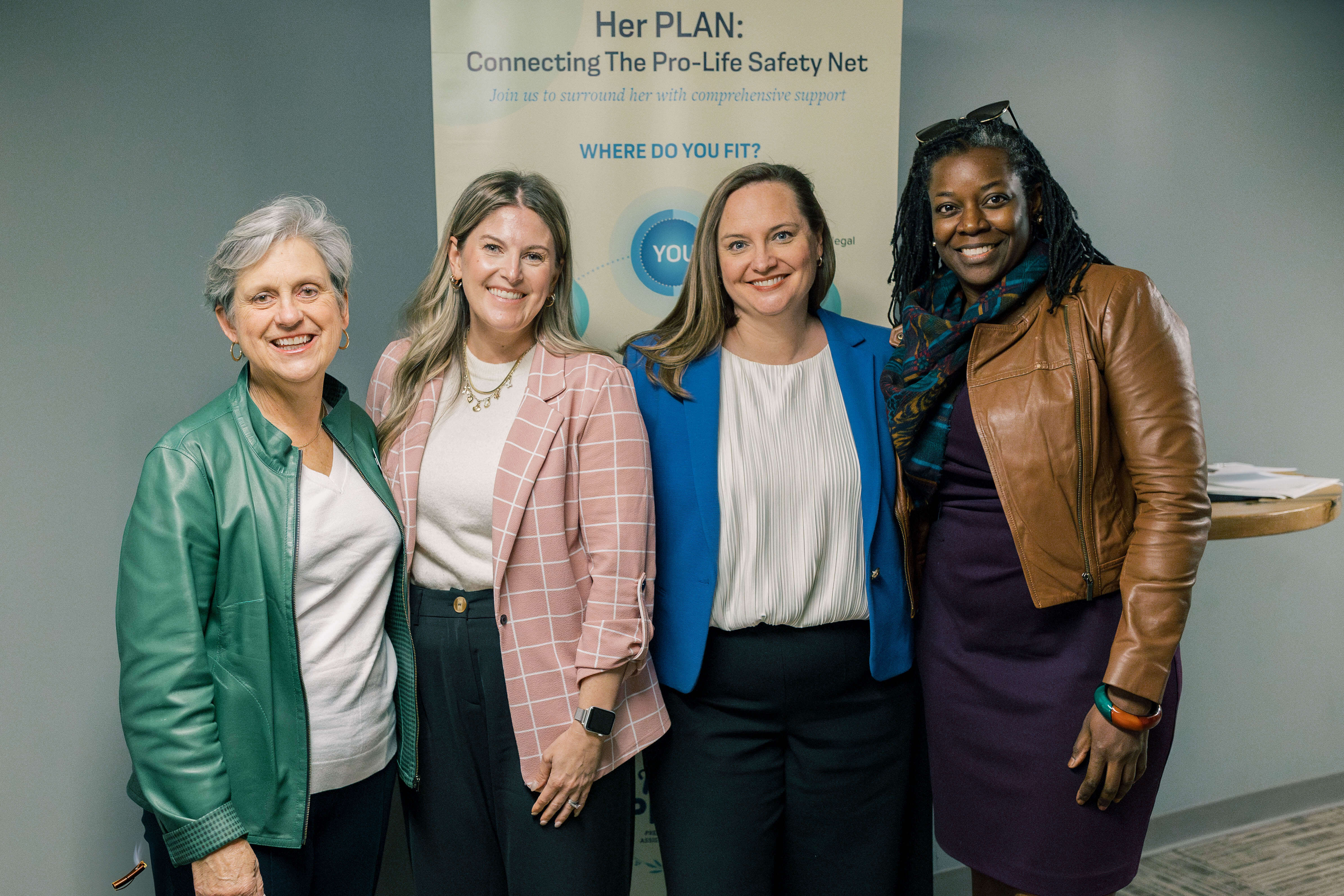 Four women stand together smiling at the camera in front of a banner that reads Her PLAN: Connecting The Pro-Life Safety Net.” They are dressed in business casual attire and appear to be at an event or gathering.