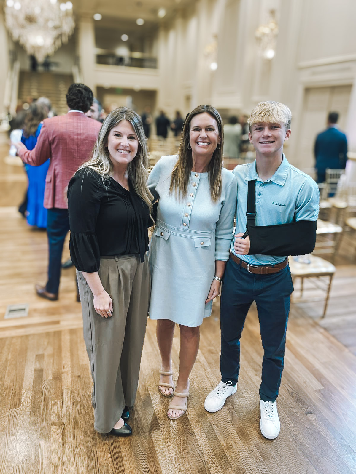 Three people stand smiling indoors at a formal event. Two women are on the left, one in a black top and khaki pants, the other in a light blue dress. A young man with a sling on his right arm stands on the right, wearing a blue polo and dark pants.