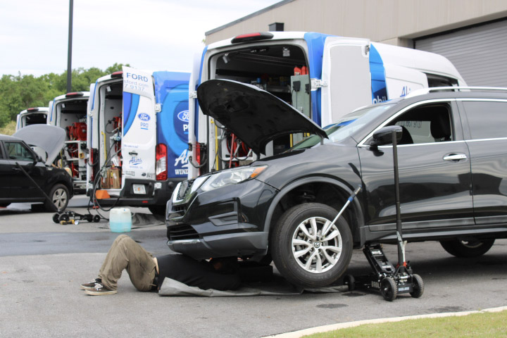 A mechanic lies under a black SUV with its hood open and a jack lifting the front, while two service vans with open doors are parked nearby. Tools and equipment are visible around the area.