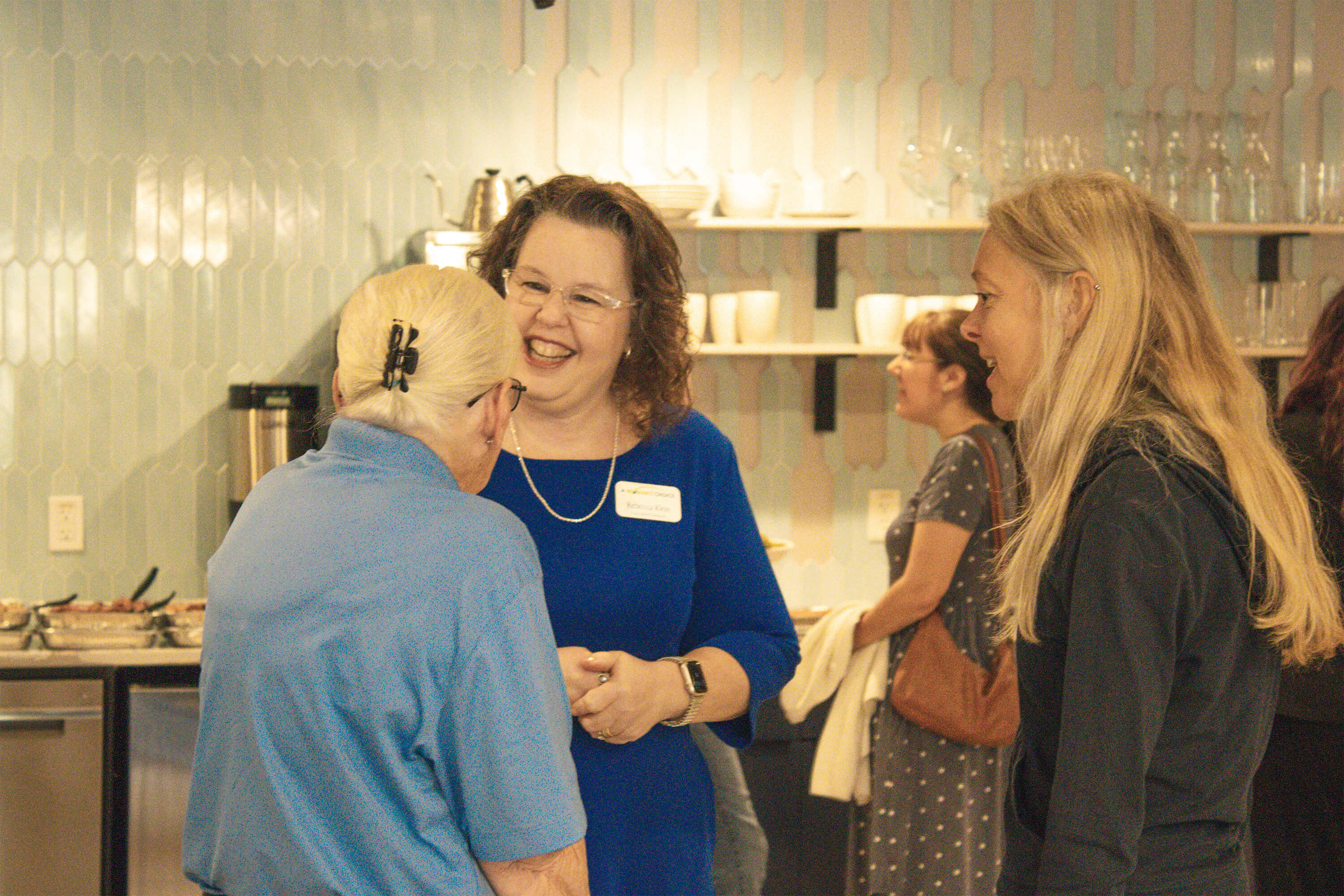 Three women stand and talk, smiling and laughing in a kitchen setting. Other people are visible in the background near shelves with dishes and glassware. The atmosphere appears friendly and casual.