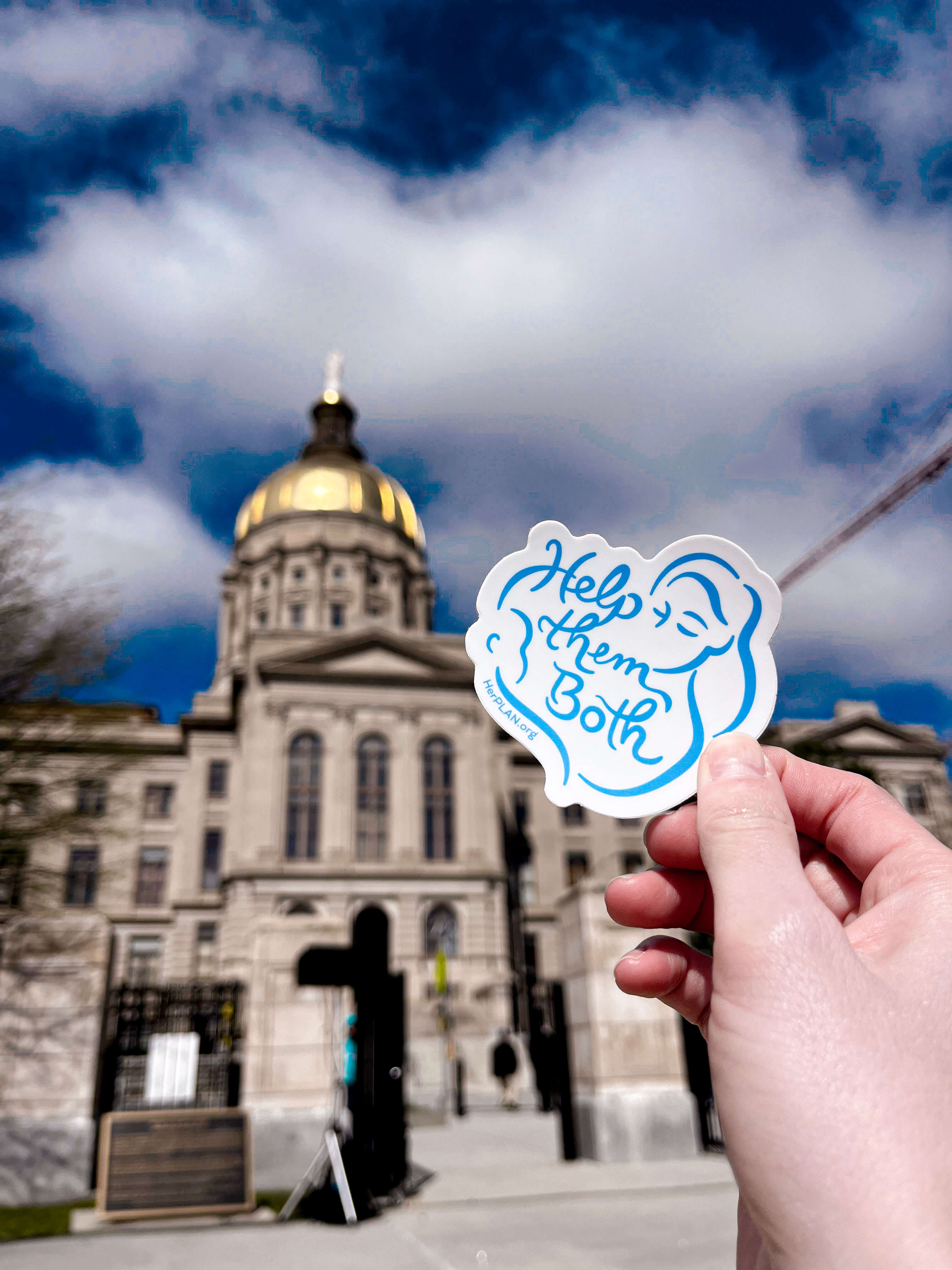A hand holds a sticker with the text Help Them Both and an illustration of a woman and baby, in front of a domed government building under a partly cloudy sky.