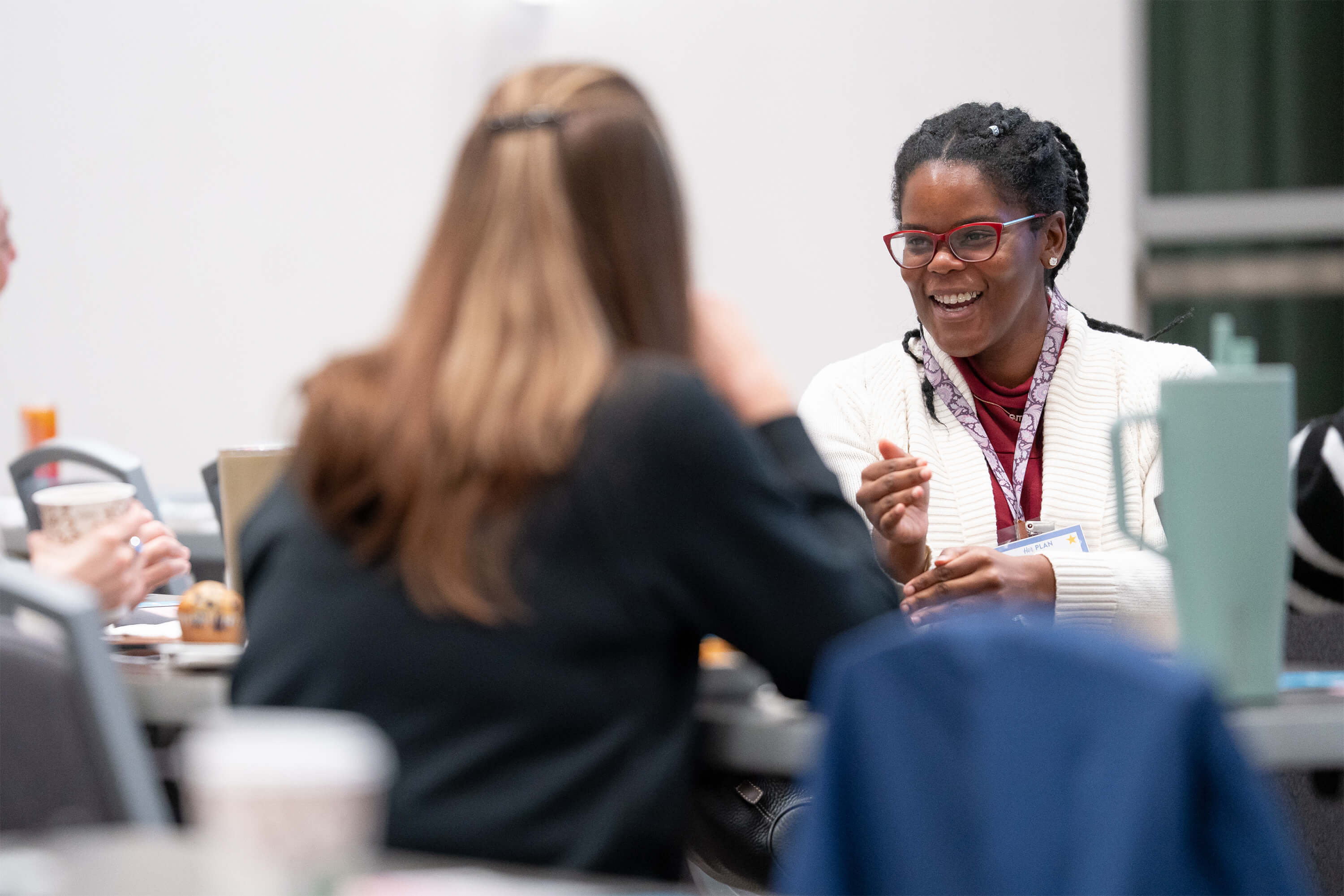 Two women sit at a table in conversation; one faces the camera smiling and wearing red glasses and a white sweater, while the other is seen from behind. Other people and objects are visible in the blurred background.