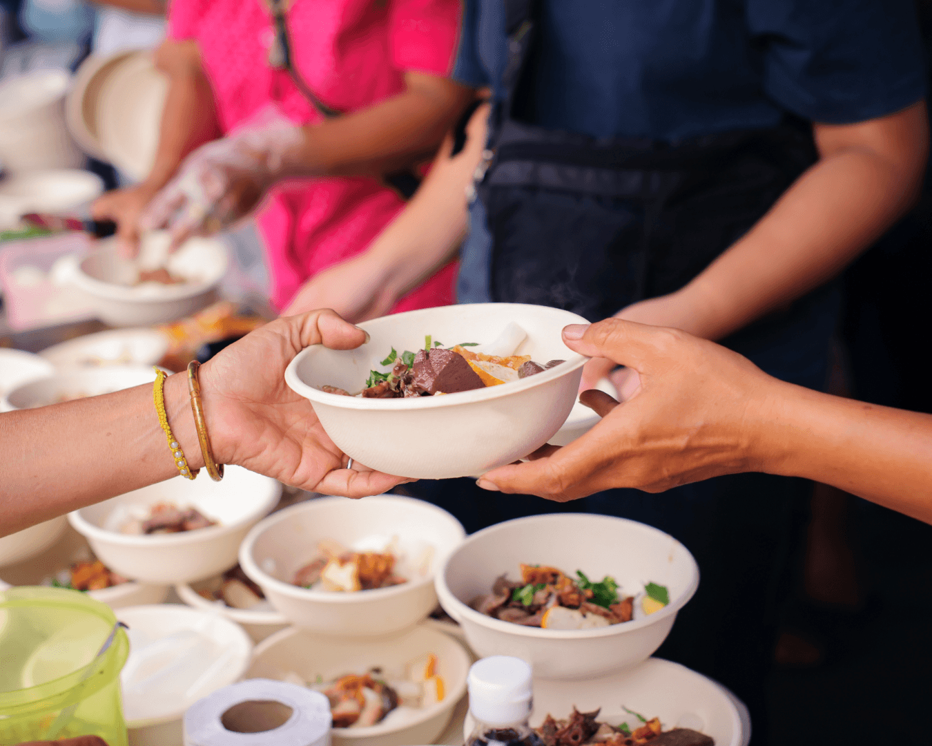 A person hands a bowl of food to another at an outdoor event, with multiple bowls of food and people in the background.