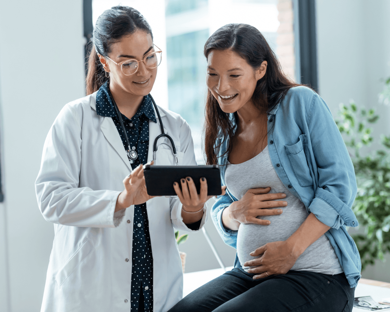 A doctor in a white coat shows a tablet to a smiling pregnant woman who is touching her stomach. They are in a bright medical office with plants in the background.