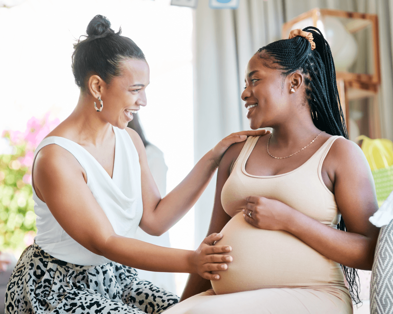 Two women smiling together; one is pregnant and wearing a beige tank top while the other, in a white sleeveless blouse, gently touches her pregnant belly in a supportive, joyful moment indoors.