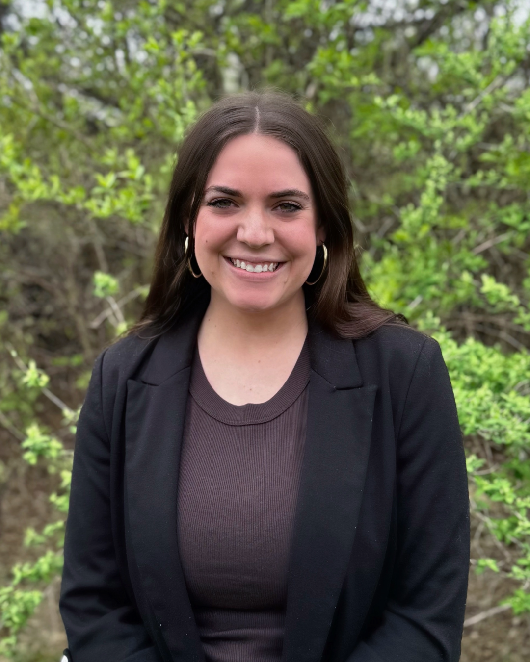 A woman with long brown hair, wearing a black blazer and brown top, smiles while standing outdoors in front of green leafy bushes.