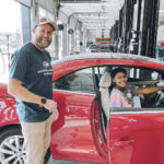 A man stands next to a red car with the drivers door open, smiling at the camera. A woman sits in the drivers seat, also smiling. They appear to be inside an automotive service garage.