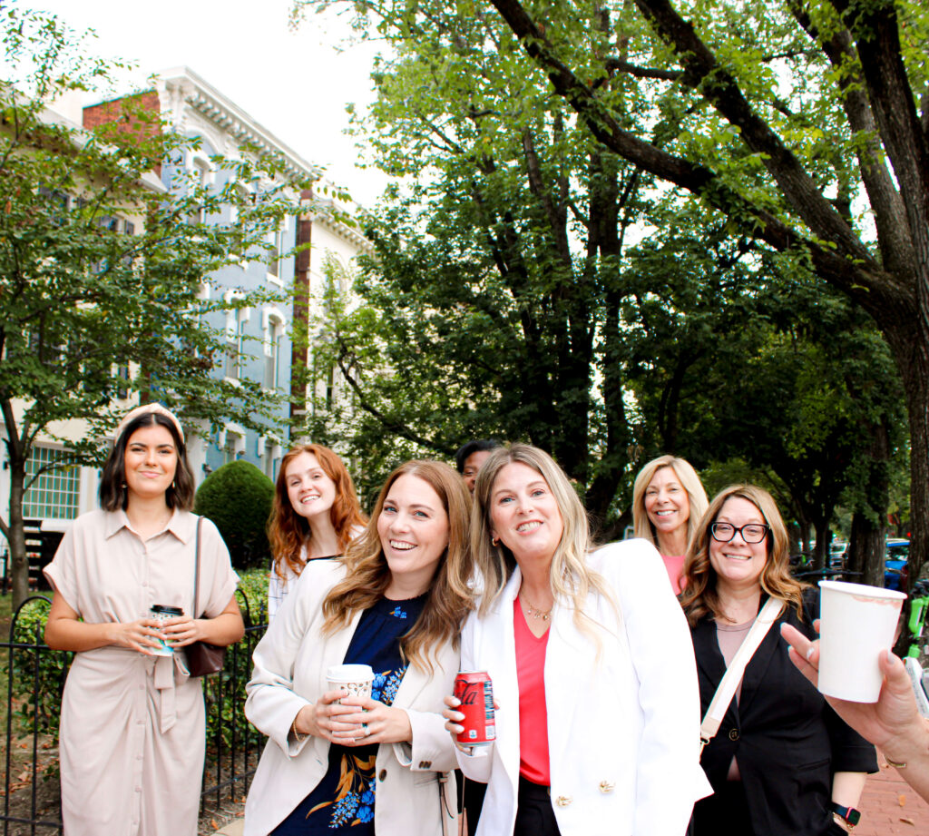 A group of seven women stand outdoors, smiling and holding drinks, with trees and buildings in the background on a bright day.